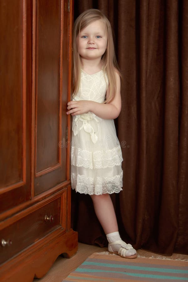 Lovely little round-faced girl with long, blonde hair below the shoulders, in a white dress. The girl peeks out from behind the vintage wardrobe. Lovely little round-faced girl with long, blonde hair below the shoulders, in a white dress. The girl peeks out from behind the vintage wardrobe