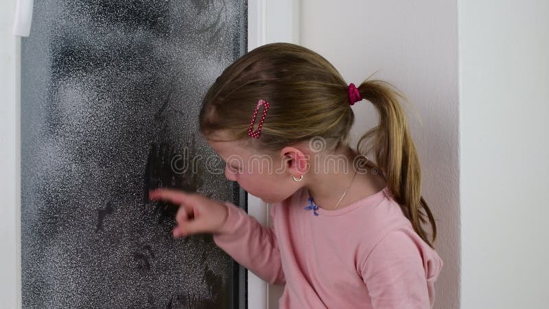 Das Mädchen zeichnet einen Finger auf einer Fensterscheibe Nettes kleines Mädchen wischt gefrorenes Fenster in der Winterzeit ab stock video footage Das Mädchen zeichnet einen Finger auf einer Fensterscheibe Nettes kleines Mädchen wischt gefrorenes Fenster in der Winterzeit ab stock video footage