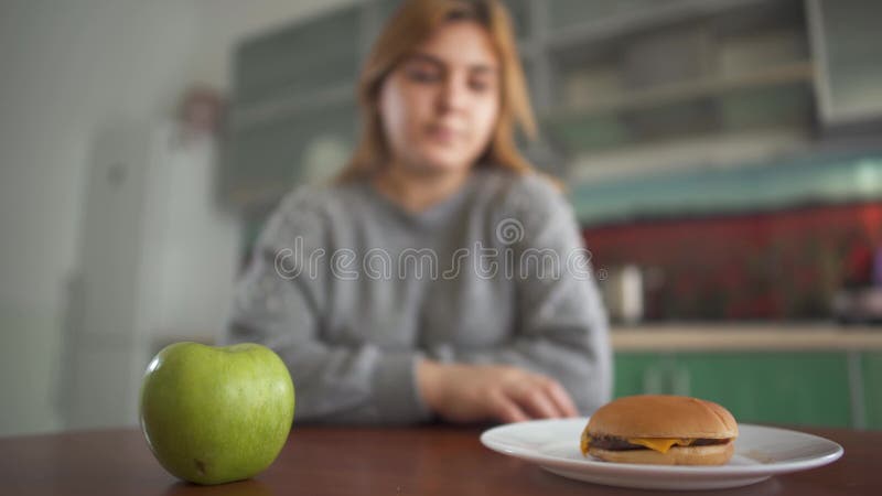 Das Plusgrößenmädchen, das auf einem unscharfen Hintergrund sitzt, denkt, dass sie einen geschmackvollen Hamburger oder einen saf