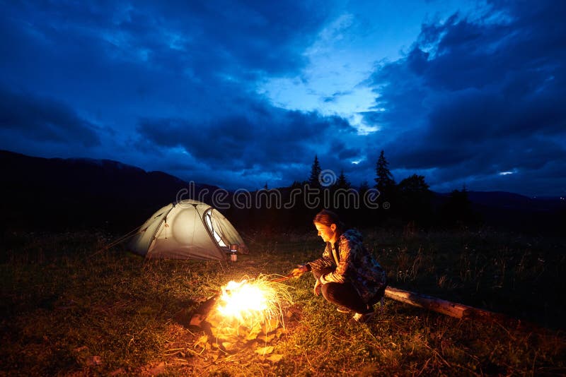 Das touristische Stillstehen der Frau nachts kampierend in den Bergen nähern sich Lagerfeuer und Zelt unter der Glättung des bewö lizenzfreie stockfotos Das touristische Stillstehen der Frau nachts kampierend in den Bergen nähern sich Lagerfeuer und Zelt unter der Glättung des bewö lizenzfreie stockfotos