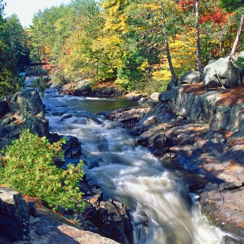 Ruggenti offuscata goccia d'acqua e le rocce di Daves cascate del niagara, vicino a Amberg wisconsin, in autunno, con la caduta del colore che mostra in Acero e pioppo e betulla alberi intorno a un piccolo ponte pedonale a distanza nel nord del nord del wisconsin, negli stati uniti d'america ed è tipica scena di foreste dello stato del nord che è seduto sul robusto scarpata del niagara 