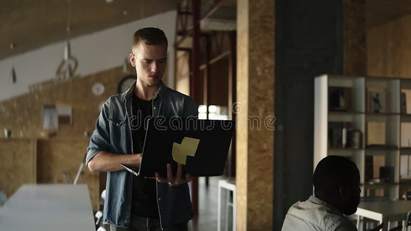 De aantrekkelijke geconcentreerde jonge bedrijfsmens loopt met laptop in zijn hand en het typen Kaukasische mens die binnen werke