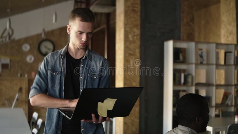 De aantrekkelijke geconcentreerde jonge bedrijfsmens loopt met laptop in zijn hand en het typen Kaukasische mens die binnen werke