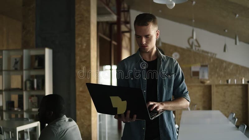 De aantrekkelijke geconcentreerde jonge bedrijfsmens loopt met laptop in zijn hand en het typen Kaukasische positieve mens