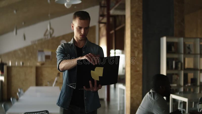 De aantrekkelijke geconcentreerde jonge bedrijfsmens loopt met zwarte laptop in zijn hand en het typen Kaukasische mens die binne