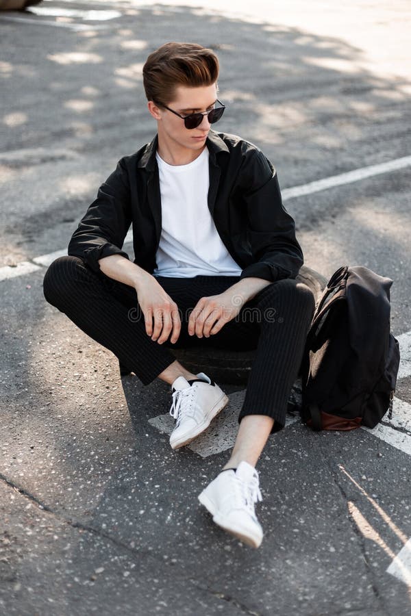 De aantrekkelijke jonge mens in een elegant overhemd in modieuze zonnebril in broek met een rugzak in in tennisschoenen zit op he stock afbeelding De aantrekkelijke jonge mens in een elegant overhemd in modieuze zonnebril in broek met een rugzak in in tennisschoenen zit op he stock afbeelding