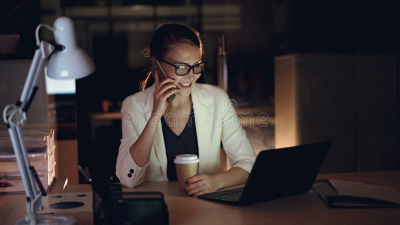 De aantrekkelijke vrouwelijke ondernemer spreekt op mobiele telefoon en gebruikt laptop die in bureau laat bij nacht werken moder