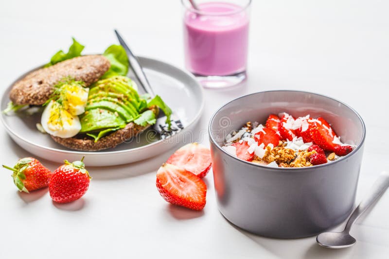 De aardbei smoothie kom met granola, de kokosnoot, de bessen en de avocado roosteren op een witte achtergrond veganistontbijt royalty-vrije stock foto De aardbei smoothie kom met granola, de kokosnoot, de bessen en de avocado roosteren op een witte achtergrond veganistontbijt royalty-vrije stock foto