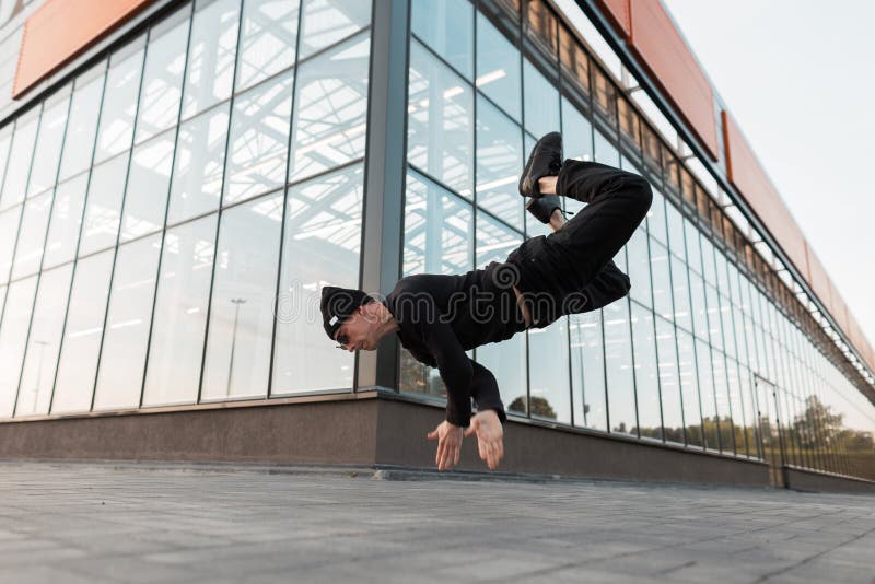 De atletische jonge mens in zwarte kleren in een modieuze hoed stelt in openlucht in de lucht dichtbij een glas modern gebouw Mod stock afbeeldingen De atletische jonge mens in zwarte kleren in een modieuze hoed stelt in openlucht in de lucht dichtbij een glas modern gebouw Mod stock afbeeldingen