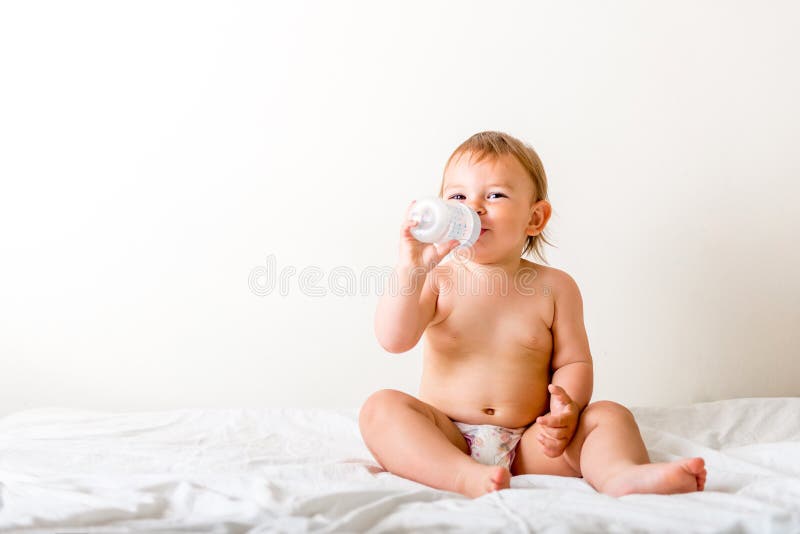 De babypeuter zit op het witte bed, glimlachen en drinkt water van plastic fles De ruimte van het exemplaar royalty-vrije stock foto's De babypeuter zit op het witte bed, glimlachen en drinkt water van plastic fles De ruimte van het exemplaar royalty-vrije stock foto's