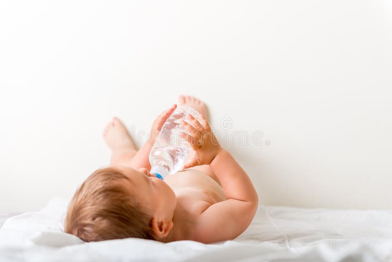 De babypeuter zit op het witte bed, glimlachen en drinkt water van plastic fles De ruimte van het exemplaar stock foto's De babypeuter zit op het witte bed, glimlachen en drinkt water van plastic fles De ruimte van het exemplaar stock foto's
