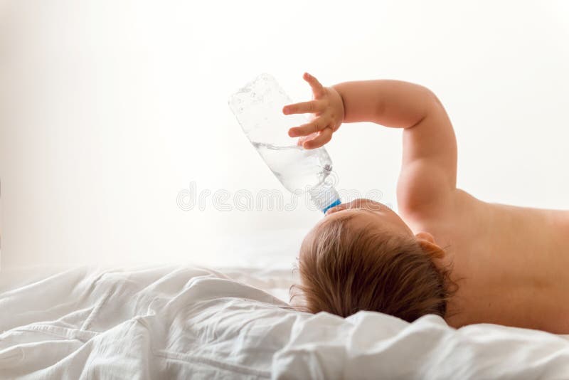 De babypeuter zit op het witte bed, glimlachen en drinkt water van plastic fles De ruimte van het exemplaar royalty-vrije stock afbeeldingen De babypeuter zit op het witte bed, glimlachen en drinkt water van plastic fles De ruimte van het exemplaar royalty-vrije stock afbeeldingen