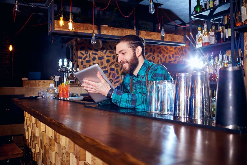 De barman met een hipsterbaard bekijkt binnen laptop achter teller stock fotografie De barman met een hipsterbaard bekijkt binnen laptop achter teller stock fotografie