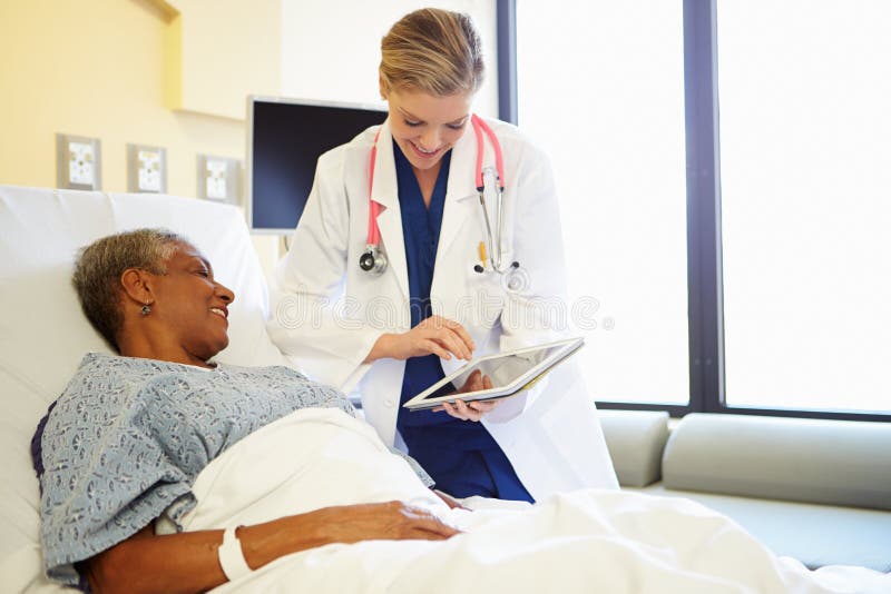 Doctor With Digital Tablet Talks To Woman In Hospital Bed Whilst Looking At Screen Smiling. Doctor With Digital Tablet Talks To Woman In Hospital Bed Whilst Looking At Screen Smiling