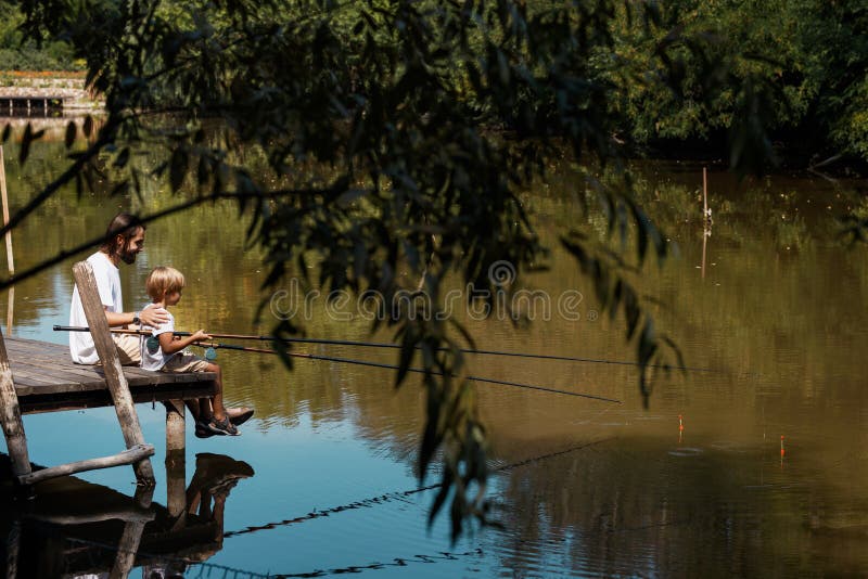 De bygdnaturen, träden och sjön i de gröna färgerna Fader och son som sitter på en träbro och ett väntande på a arkivbilder De bygdnaturen, träden och sjön i de gröna färgerna Fader och son som sitter på en träbro och ett väntande på a arkivbilder