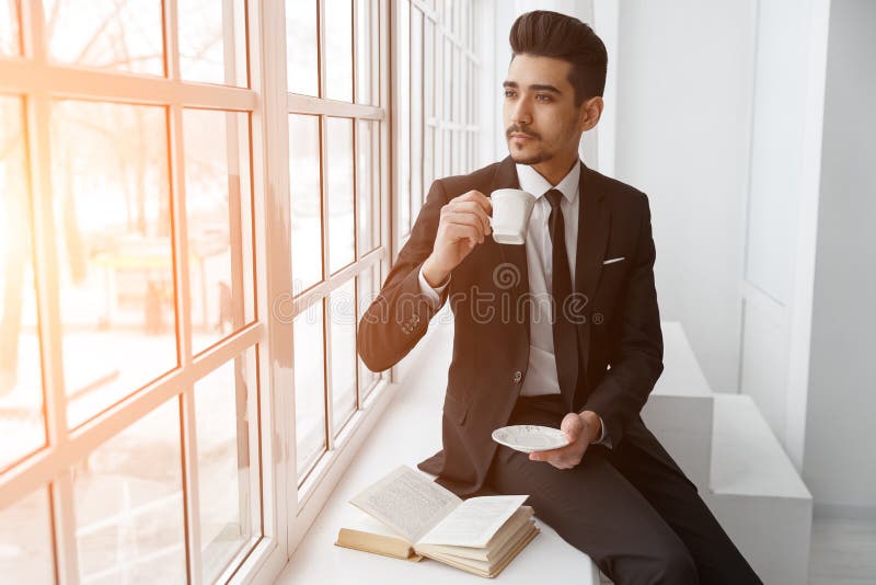 De denkende zakenman in kostuumzitting op vensterbank en drinkt koffie stock afbeelding De denkende zakenman in kostuumzitting op vensterbank en drinkt koffie stock afbeelding