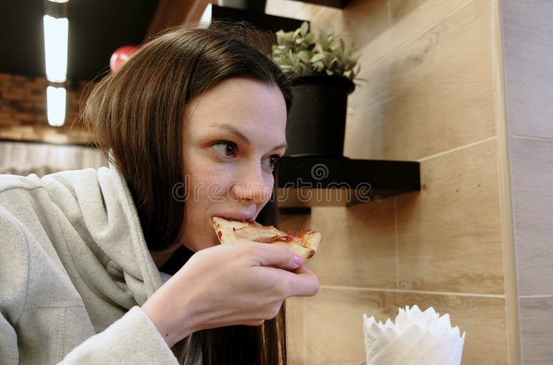 De donkerbruine jonge hongerige vrouwenbeten van een smakelijke plak van pizza en eten het, zijaanzicht stock fotografie De donkerbruine jonge hongerige vrouwenbeten van een smakelijke plak van pizza en eten het, zijaanzicht stock fotografie