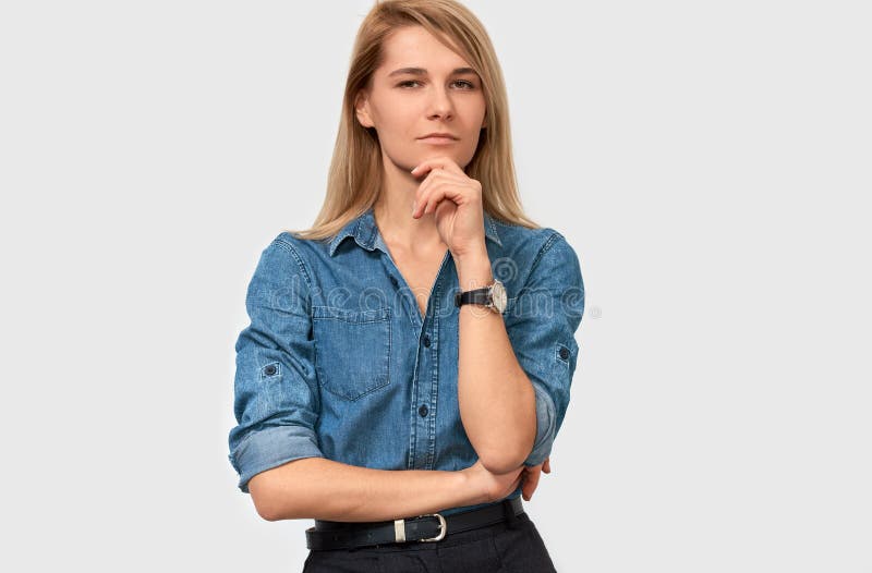 De ernstige mooie blonde jonge vrouw die denimoverhemd met vinger dragen vouwde op de kin die haar gezicht fronsen en aan de came stock foto's De ernstige mooie blonde jonge vrouw die denimoverhemd met vinger dragen vouwde op de kin die haar gezicht fronsen en aan de came stock foto's