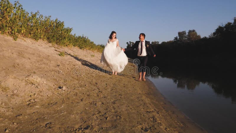 De gelukkige bruid en de bruidegom lopen blootvoets op strand langs de rivier