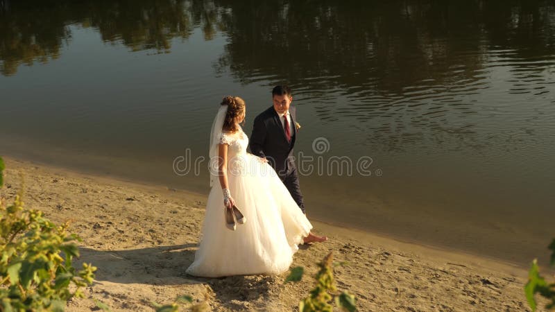 De gelukkige bruid in witte kleding en de bruidegom in kostuum lopen blootvoets op zand langs rand van riverbank het houdende van