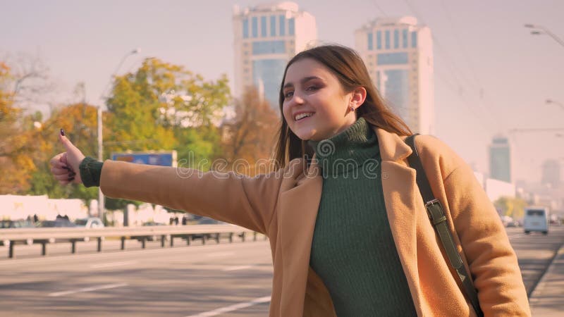 De gelukkige leuke Kaukasische modieuze vrouw lift op weg in zonnige dag met weg en carr op achtergrond