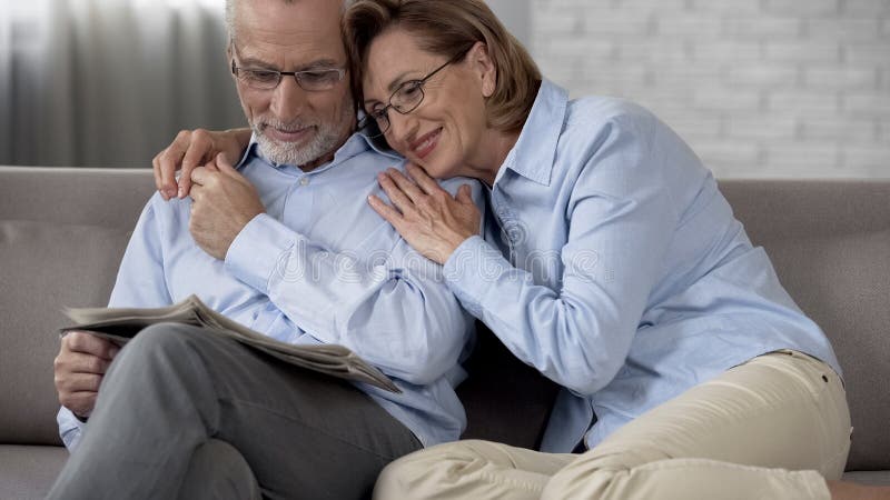 De gelukkige zitting van het gepensioneerdenpaar op laag, de krant die van de mensenlezing, dame hem koesteren stock fotografie De gelukkige zitting van het gepensioneerdenpaar op laag, de krant die van de mensenlezing, dame hem koesteren stock fotografie