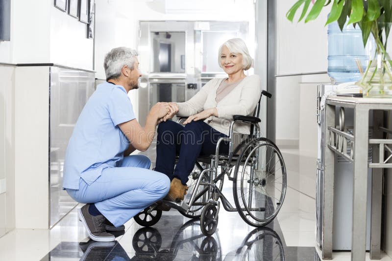 Mature male physiotherapist holding smiling senior woman's hand on wheelchair in rehab center. Mature male physiotherapist holding smiling senior woman's hand on wheelchair in rehab center