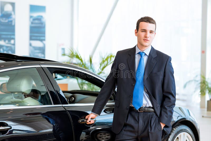 Dealer stands near a new car in the showroom, put one hand on the car. Dealer stands near a new car in the showroom, put one hand on the car