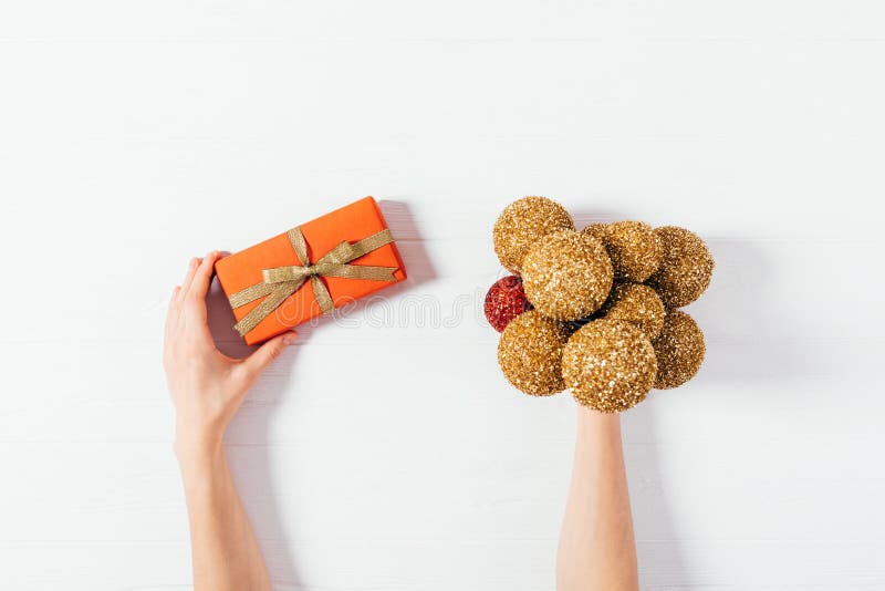 De handen die van het wijfje rode giftdoos met lint en boog naast bos van glanzende gouden Kerstmisballen houden op witte achterg stock foto De handen die van het wijfje rode giftdoos met lint en boog naast bos van glanzende gouden Kerstmisballen houden op witte achterg stock foto
