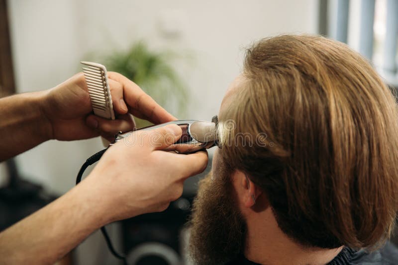 De handen van jonge kapper die tot kapsel maken aan de aantrekkelijke gebaarde mens in herenkapper stock afbeelding De handen van jonge kapper die tot kapsel maken aan de aantrekkelijke gebaarde mens in herenkapper stock afbeelding