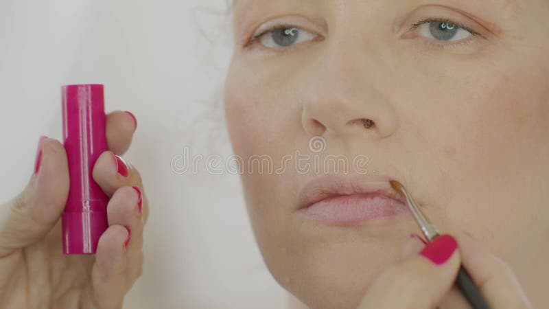 De handen van de make - up kunstenaar passen lippenstift toe met een borstel op de lippen die het gezicht van een blonde caucasiaa