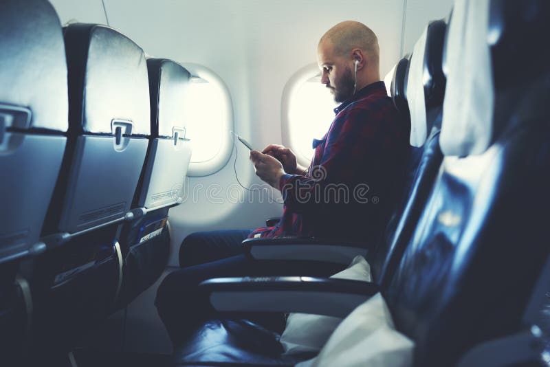 De Hipsterkerel let op video op celtelefoon, terwijl in vliegtuig zit royalty-vrije stock fotografie De Hipsterkerel let op video op celtelefoon, terwijl in vliegtuig zit royalty-vrije stock fotografie