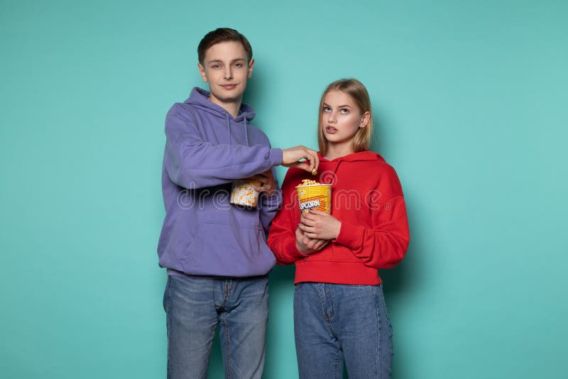 De jonge aantrekkelijke mens in blauwe hoodie stealing popcorn van zijn meisje, mooi blondemeisje bekijkt verontwaardigd hem stock fotografie De jonge aantrekkelijke mens in blauwe hoodie stealing popcorn van zijn meisje, mooi blondemeisje bekijkt verontwaardigd hem stock fotografie