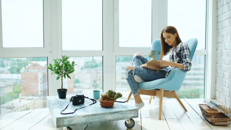 De jonge aantrekkelijke vrouw las boek en drinkt koffiezitting op balkon in moderne zolderflat stock afbeeldingen De jonge aantrekkelijke vrouw las boek en drinkt koffiezitting op balkon in moderne zolderflat stock afbeeldingen