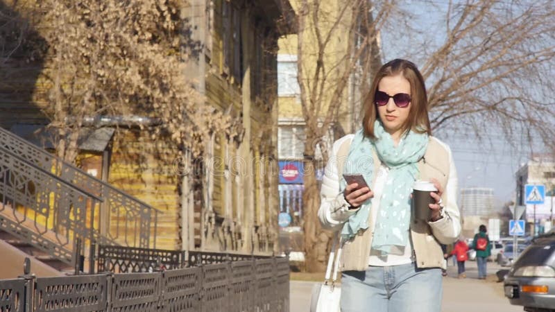 De jonge bedrijfsvrouw loopt op de straat en drinkt koffie en het gebruiken van smartphone bij middagpauze stock video De jonge bedrijfsvrouw loopt op de straat en drinkt koffie en het gebruiken van smartphone bij middagpauze stock video