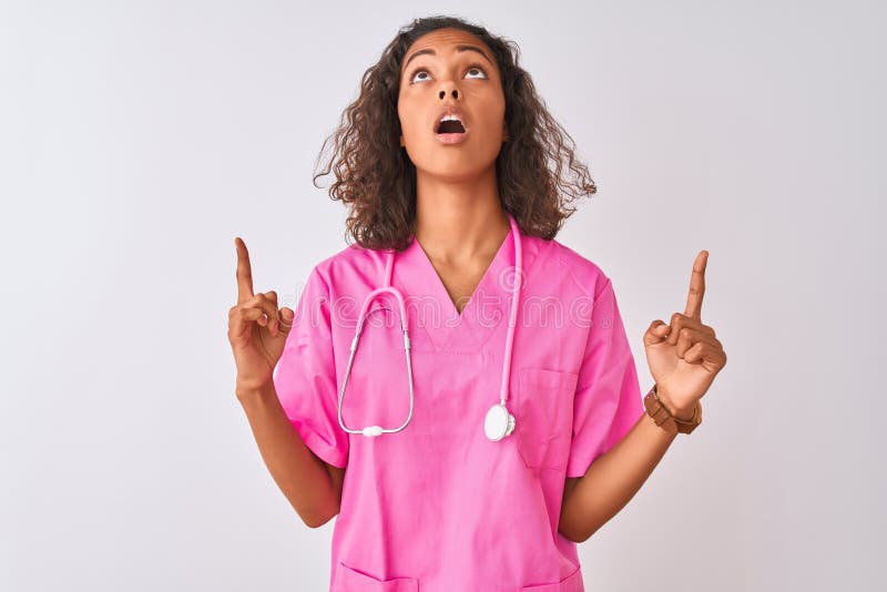 Young brazilian nurse woman wearing stethoscope standing over isolated white background amazed and surprised looking up and pointing with fingers and raised arms. Young brazilian nurse woman wearing stethoscope standing over isolated white background amazed and surprised looking up and pointing with fingers and raised arms