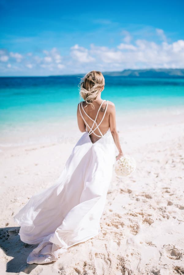 Young blonde long hair bride in long white dress standing on the white sand beach. Tropical turquois ocean on the background. Young blonde long hair bride in long white dress standing on the white sand beach. Tropical turquois ocean on the background