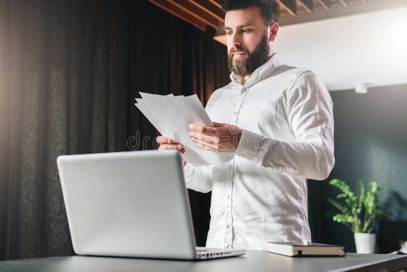 De jonge gebaarde atrractive zakenman in wit overhemd bevindt zich dichtbij bureau voor laptop, lezend documenten stock afbeelding De jonge gebaarde atrractive zakenman in wit overhemd bevindt zich dichtbij bureau voor laptop, lezend documenten stock afbeelding