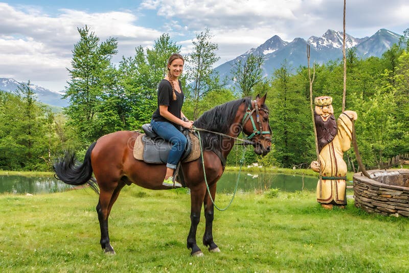 De jonge gelukkige glimlachende Kaukasische vrouw die jeans dragen berijdt bruin paard die en bij camera in de bergplatteland van royalty-vrije stock foto De jonge gelukkige glimlachende Kaukasische vrouw die jeans dragen berijdt bruin paard die en bij camera in de bergplatteland van royalty-vrije stock foto