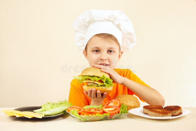 De jonge grappige jongen in chef-kokshoed geniet van kokend smakelijke hamburger
