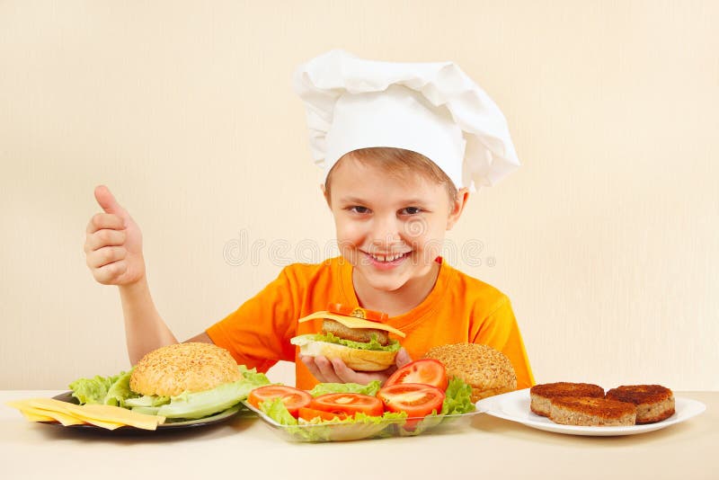 De jonge grappige jongen in chef-kokshoed toont hoe te om hamburger te koken royalty-vrije stock foto De jonge grappige jongen in chef-kokshoed toont hoe te om hamburger te koken royalty-vrije stock foto