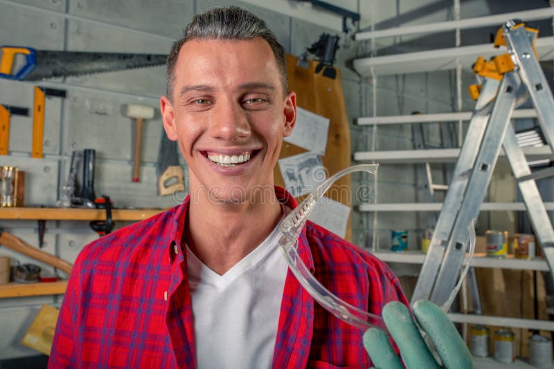 De jonge knappe glimlachende Kaukasische timmerman houdt de veiligheidsbril in van hem zijn workshop indient stock afbeelding De jonge knappe glimlachende Kaukasische timmerman houdt de veiligheidsbril in van hem zijn workshop indient stock afbeelding