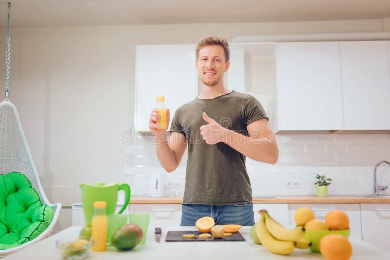 De jonge knappe mens houdt duim en jus d'orange tegen terwijl het snijden van verse vruchten in de keuken Gezond voedsel vegetari royalty-vrije stock afbeeldingen De jonge knappe mens houdt duim en jus d'orange tegen terwijl het snijden van verse vruchten in de keuken Gezond voedsel vegetari royalty-vrije stock afbeeldingen