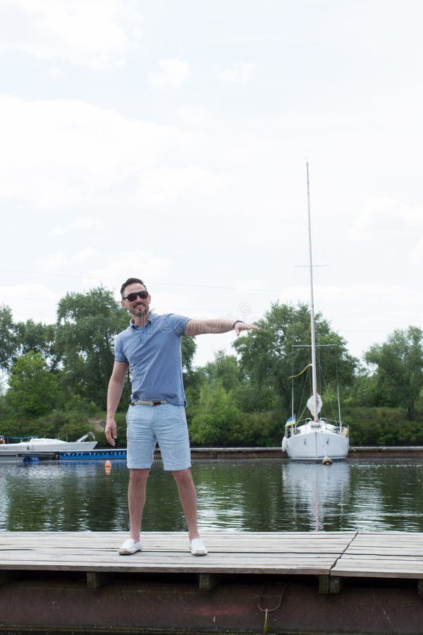 De jonge knappe zeiler die in zonglazen bij rivierpijler lopen en heet vrienden welkom De knappe zakenman nodigt aan zijn boot ui stock foto De jonge knappe zeiler die in zonglazen bij rivierpijler lopen en heet vrienden welkom De knappe zakenman nodigt aan zijn boot ui stock foto