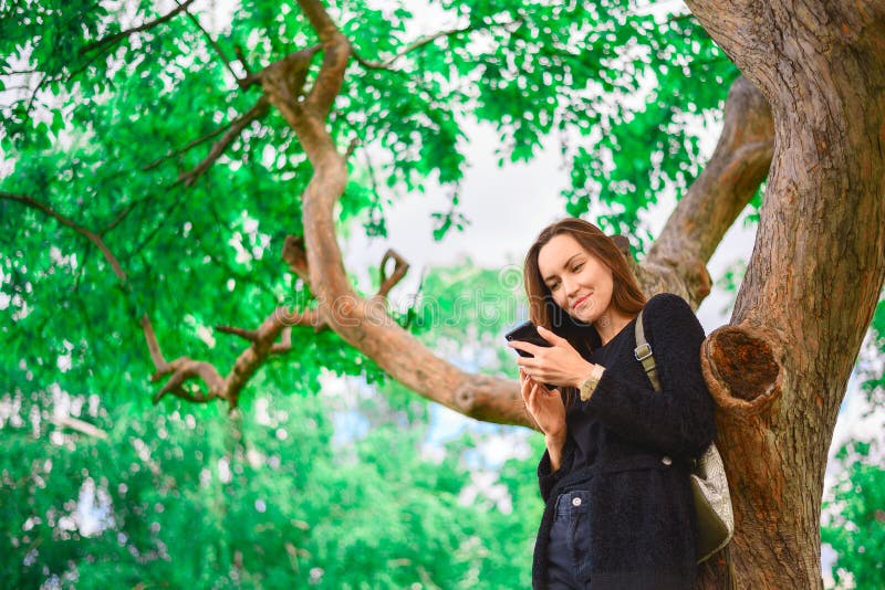 De jonge meisjestribunes bij een grote groene boom met een smartphone in haar handen, communiceert op de Internet-berichten, de m stock afbeelding De jonge meisjestribunes bij een grote groene boom met een smartphone in haar handen, communiceert op de Internet-berichten, de m stock afbeelding