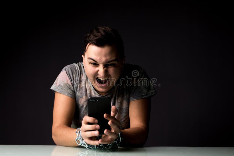De jonge Mens die aan de verslavingsschreeuw van de telefoonafhankelijkheid lijden met ketting dient donkere ruimte in stock afbeeldingen De jonge Mens die aan de verslavingsschreeuw van de telefoonafhankelijkheid lijden met ketting dient donkere ruimte in stock afbeeldingen