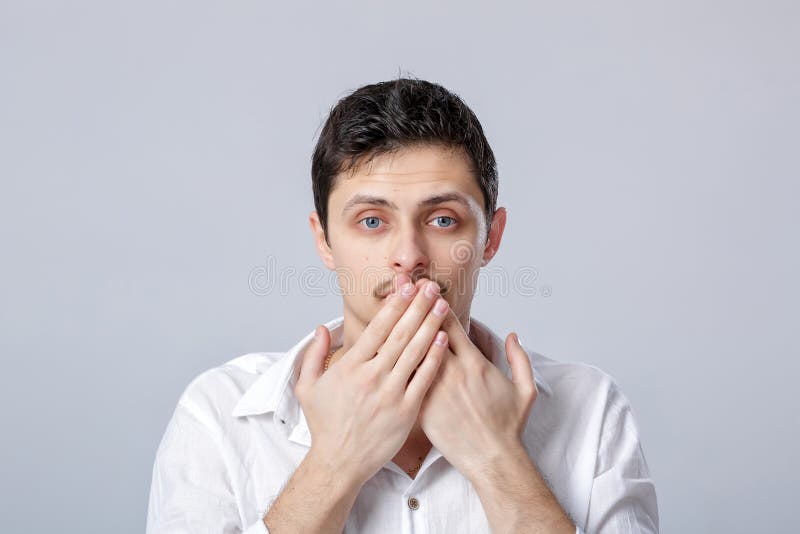De jonge mens in wit overhemd behandelt zijn mond met zijn handen op grijs stock foto De jonge mens in wit overhemd behandelt zijn mond met zijn handen op grijs stock foto