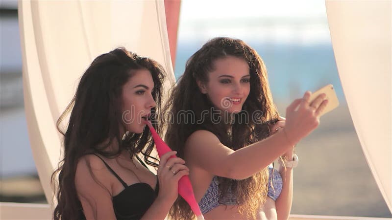 De jonge mooie sexy meisjes in bikini roken een waterpijp en maken selfie beelden op smartphone bij de bar op het strand