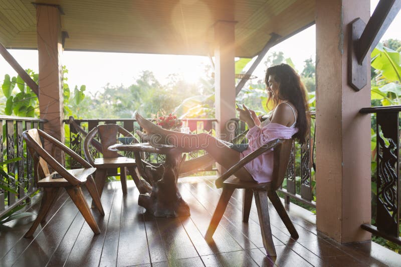 De jonge Mooie Vrouw die Cel Slimme Telefoon Sit On Terrace With Tropical Forest Landscape And Morning Sun met behulp van glanst royalty-vrije stock afbeelding De jonge Mooie Vrouw die Cel Slimme Telefoon Sit On Terrace With Tropical Forest Landscape And Morning Sun met behulp van glanst royalty-vrije stock afbeelding