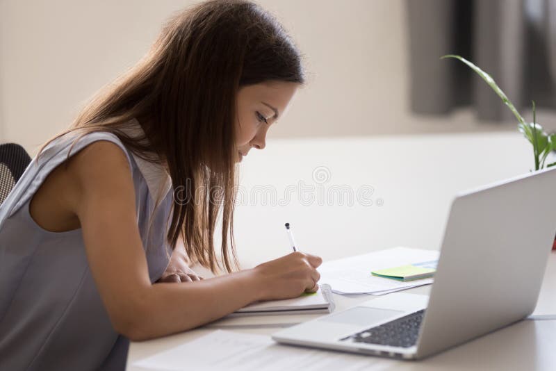 Young girl, intern holding pen in hand, making notes, working with laptop in office, female student preparing for test or exam, writing essay in notebook, studying in college, university. Young girl, intern holding pen in hand, making notes, working with laptop in office, female student preparing for test or exam, writing essay in notebook, studying in college, university